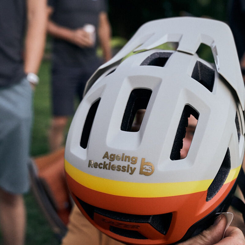 man holding up a helmet with the sticker "Ageing Recklessly" on it